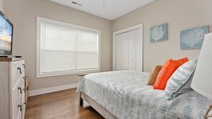 Ein Schlafzimmer mit Bett, Kommode, Fenster mit Jalousien, einer Tür und Wandbildern.