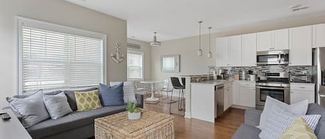 A modern living room with a grey sofa, a coffee table, and a kitchen area with white cabinets and stainless steel appliances.