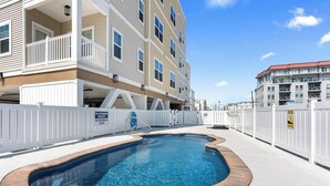 A swimming pool with a white fence, a multi-story building with balconies, and a clear blue sky. - Residence 101S at The Sandcastle Condominiums (Wildwood Crest)