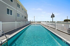 A rectangular pool with a wooden deck, surrounded by white railings, and a building with multiple windows to the left. - Oceanview 3BR Condo with Balcony & Seawall Access (North Wildwood)