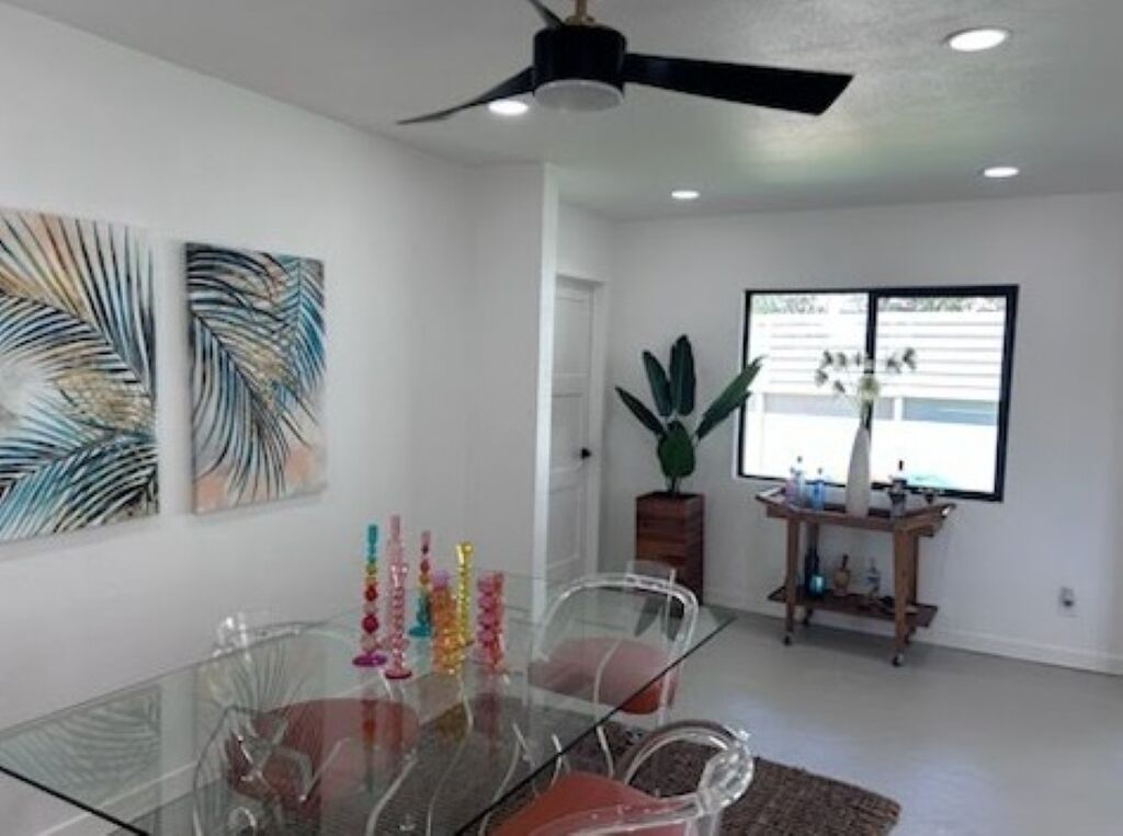 A dining area with a glass table, chairs, ceiling fan, artwork, and a window with blinds.