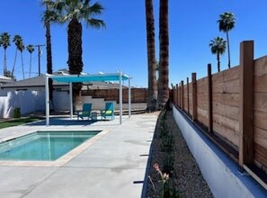 Ein Swimmingpool mit überdachter Sitzgruppe, Palmen und einem Holzzaun.