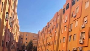 Une rue étroite en milieu urbain, bordée d’immeubles à plusieurs étages, avec une fontaine centrale et un ciel dégagé.