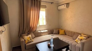 Un salon comprenant un canapé, une table basse, un téléviseur fixé au mur et une fenêtre avec des rideaux.