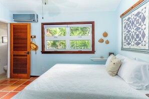 A bedroom with a bed, a window with a view of greenery, a wall-mounted air conditioner, and a wooden door. - East Gate In Coral Bay Back Up Power Included (Coral Bay)