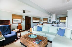 A living room with a blue armchair, a white sofa, a wicker coffee table, and a kitchen area in the background. - East Gate In Coral Bay Back Up Power Included (Coral Bay)