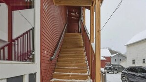 Eine Holztreppe führt zu einem Eingang im zweiten Stock, begleitet von einem Metallgeländer, roten Holzverkleidungen und einem weißen Fenster.