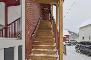 A wooden staircase leading to a second-floor entrance with a metal handrail, flanked by red siding and a white window.
