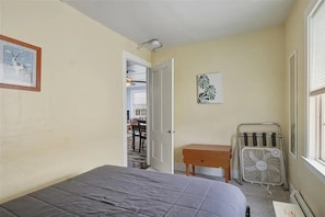 Une chambre à coucher avec un lit, une petite table, un ventilateur, un tableau accroché au mur et une porte donnant sur une autre pièce.