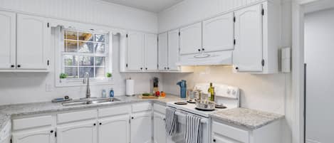 Une cuisine avec des meubles blancs, un plan de travail en granit, une cuisinière et une fenêtre avec des stores.