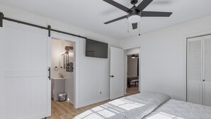 Een slaapkamer met een bed, een plafondventilator, een televisie en een badkamer met een wastafel en spiegel.