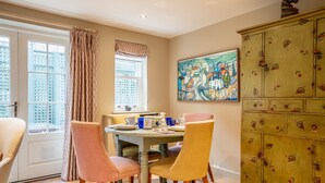 A dining area with a table set for four, chairs, a window with curtains, a painting on the wall, and a wooden cabinet with fruit designs. - St Mary’s Mews - A Charming York Retreat (York)