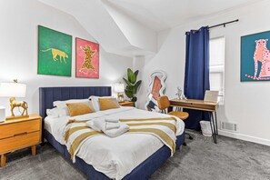 Un dormitorio con cama, mesita de noche, escritorio, silla y obras de arte en las paredes.