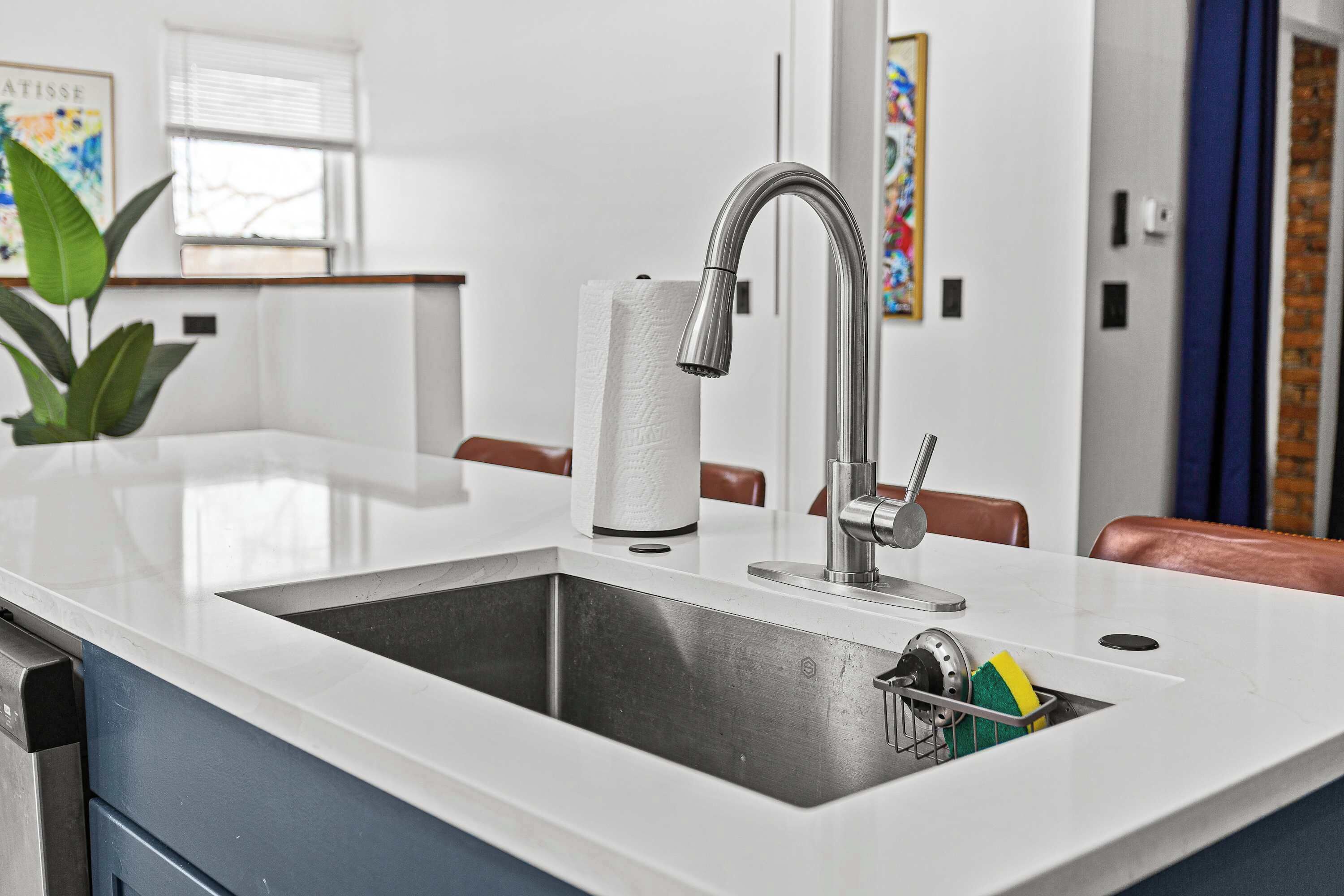 A kitchen with a stainless steel sink, a roll of paper towels, and a dish rack with sponges.