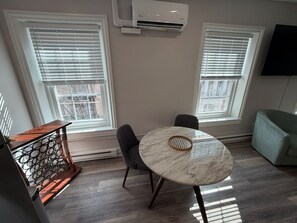 Una habitación con una mesa redonda de mármol, dos sillas grises, una mesita de noche de madera y un aire acondicionado instalado.