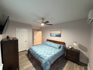 Un dormitorio con cama, ventilador de techo, mesita de noche, cómoda, puerta y un cuadro en la pared.