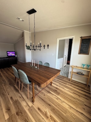 Un coin repas avec une table en bois, des chaises, un lustre et un téléviseur en arrière-plan.