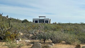 Una casa moderna de dos pisos amb teulada plana i un voladís gran al segon pis, envoltada de vegetaci ó seca i cactus.