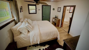 A bedroom with a bed, a ceiling fan, a window, a door, a small table, and a mirror.