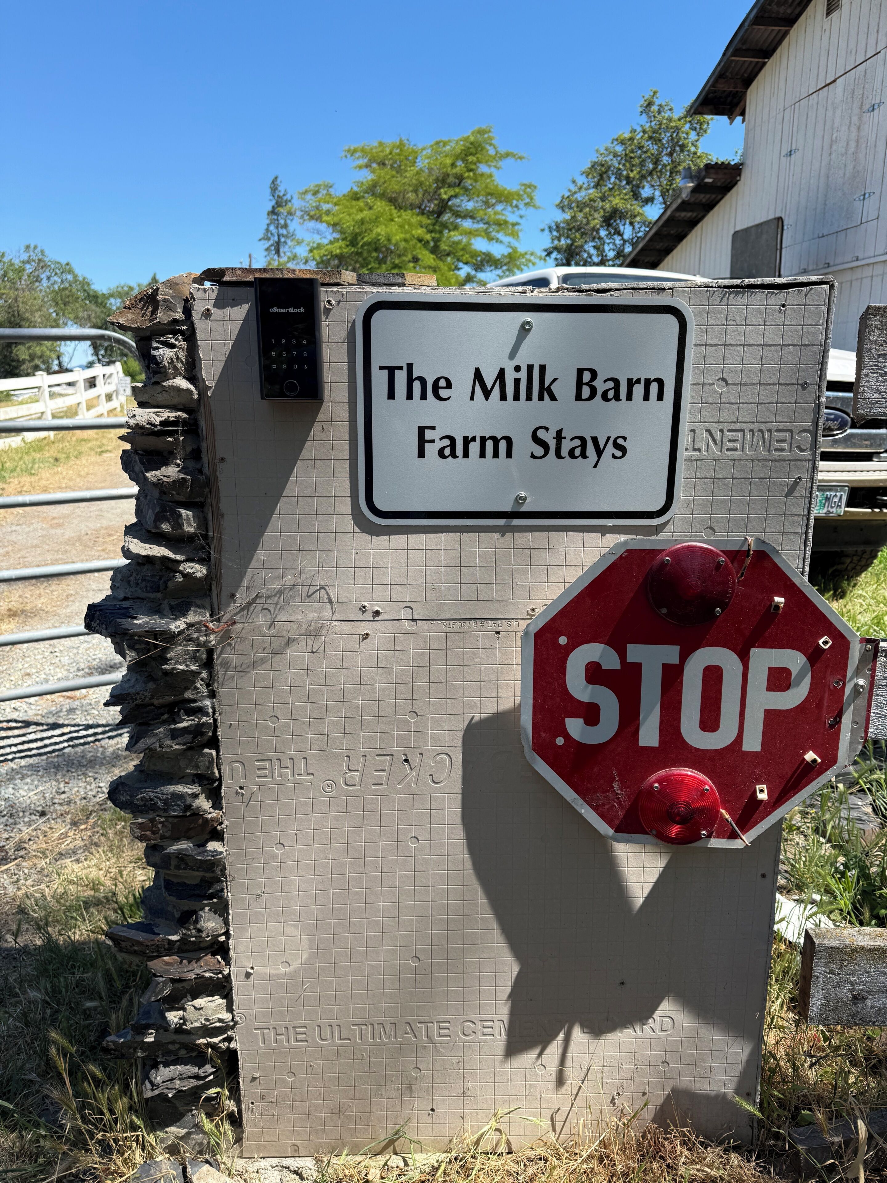 A stop sign and a sign for 'The Milk Barn Farm Stays' with a stone wall and a vehicle in the background.