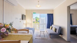 A hotel room with a bed, a sofa, a dining table with chairs, a small table with a vase of flowers, and a view of the outdoors through a window. - Claudelands Motel (Hamilton)