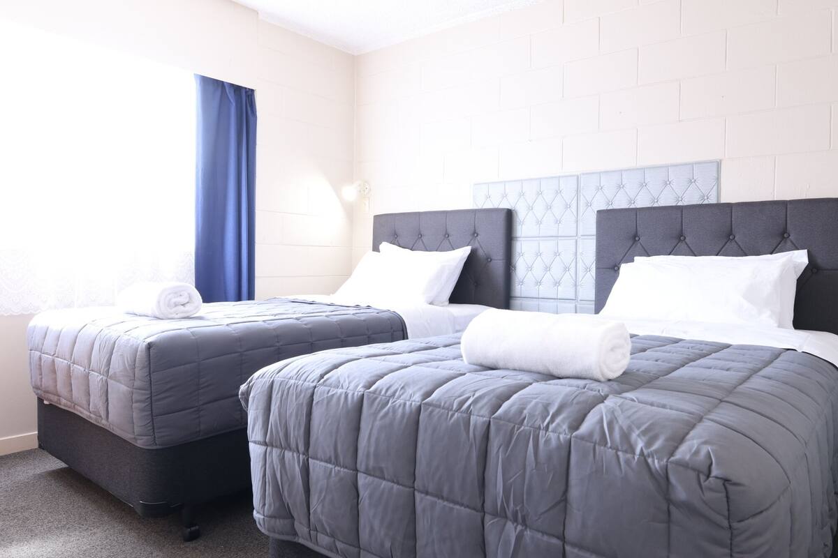 Habitación de hotel con dos camas, una ventana con cortinas azules y una pared de ladrillo blanco.