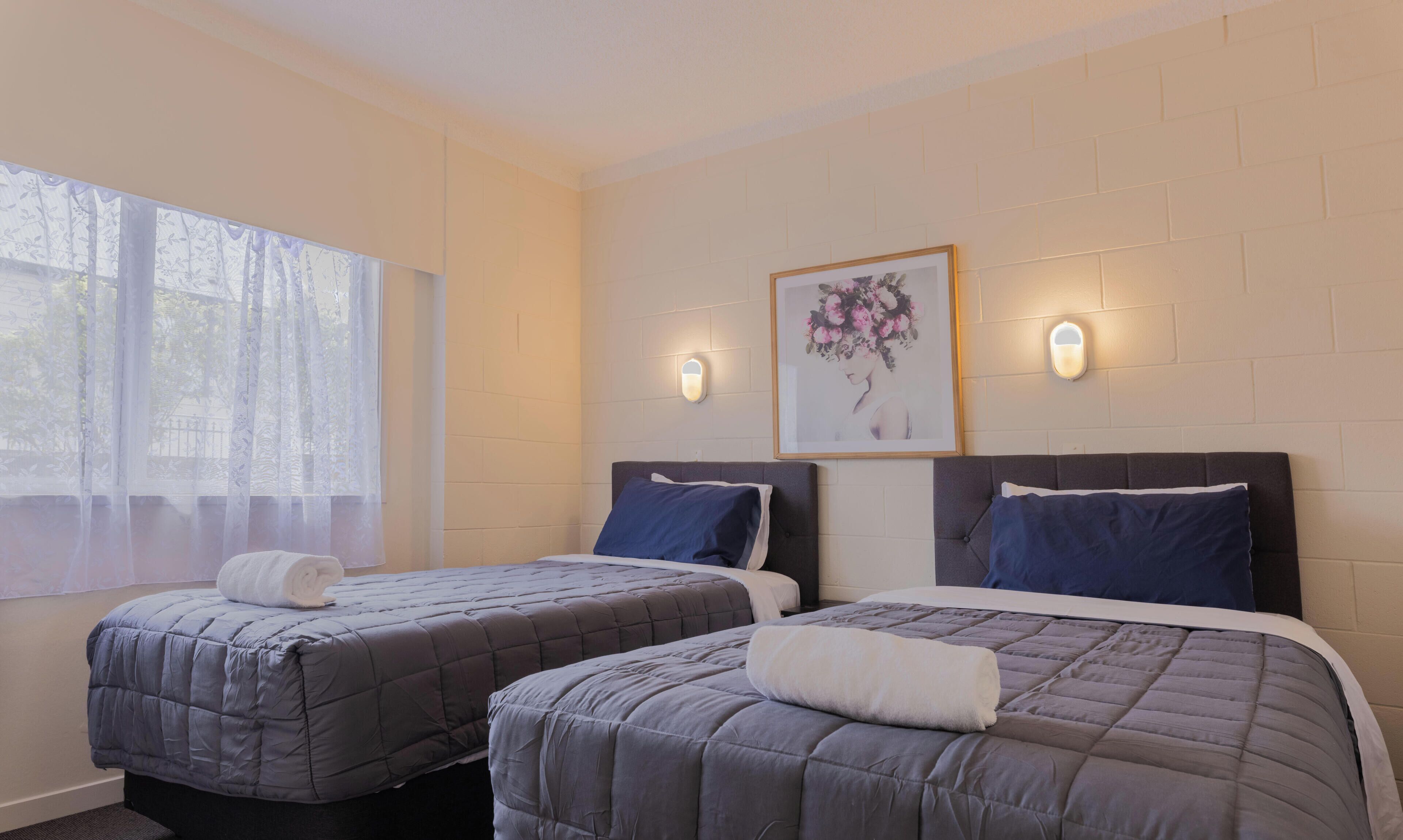A hotel room with two beds, a large window with sheer curtains, a wall-mounted picture, and two wall sconces.