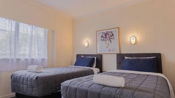 A hotel room with two beds, a large window with sheer curtains, a wall-mounted picture, and two wall sconces.