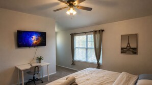 A bedroom with a bed, a desk, a ceiling fan, a window with curtains, and a wall-mounted TV displaying a galaxy image.