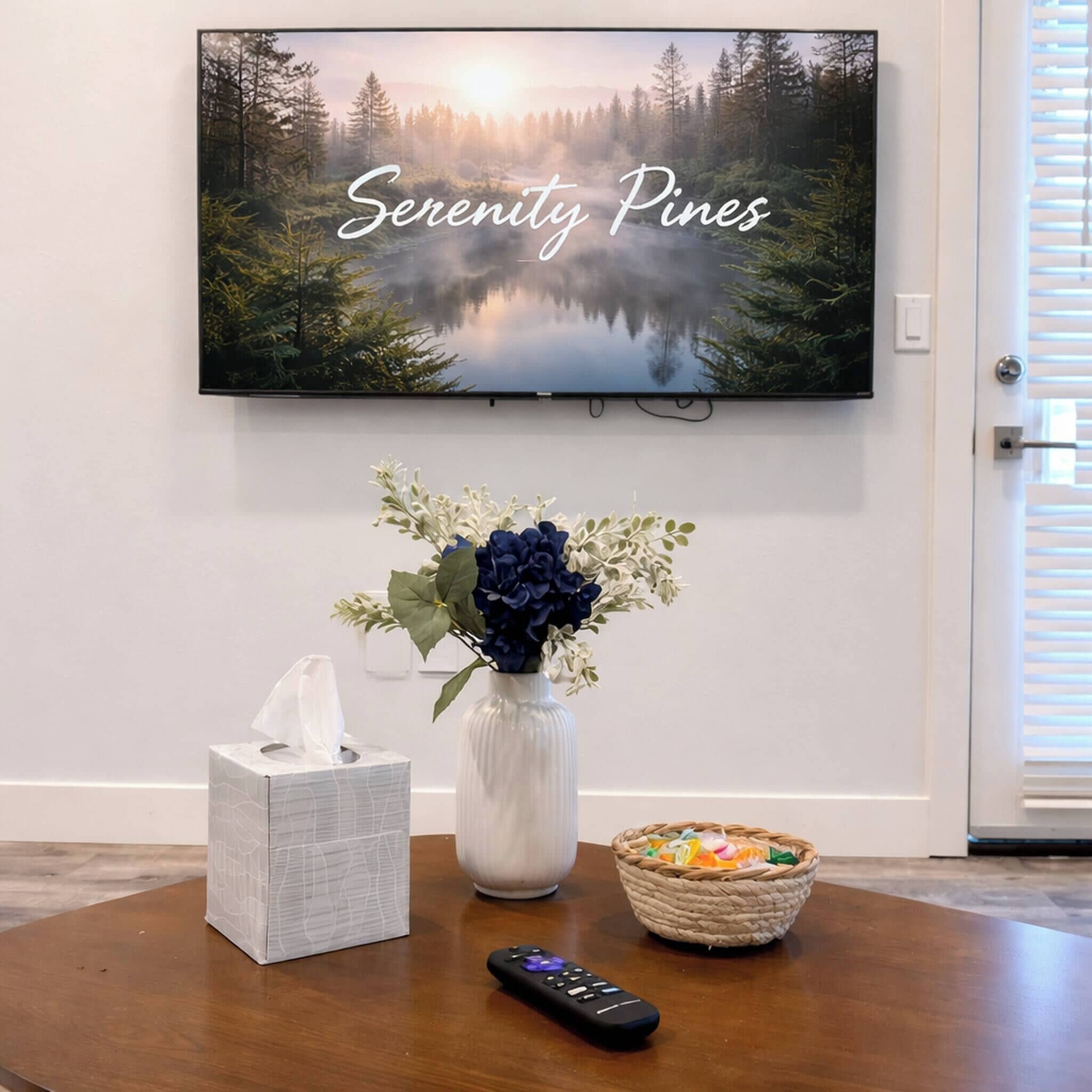 Un televisor montado en la pared mostrando 'Serenity Pines', una caja de pañuelos, un jarrón con flores, una cesta con aperitivos y un control remoto sobre una mesa de madera.