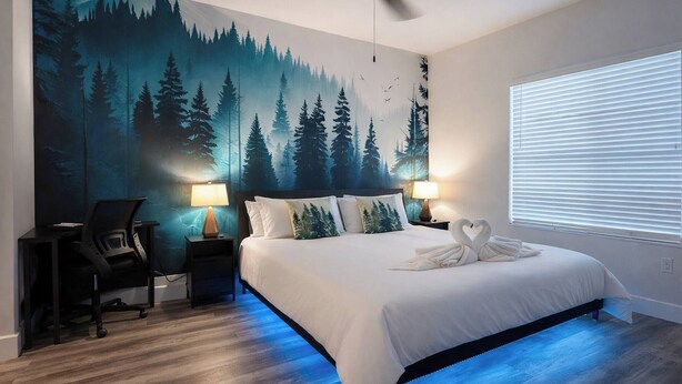 A bedroom with a bed, two bedside lamps, a desk with a chair, a ceiling fan, and a large forest mural on the wall. - Resort 1BR Oasis Pool Spa Gym Reno Getaway (Reno)
