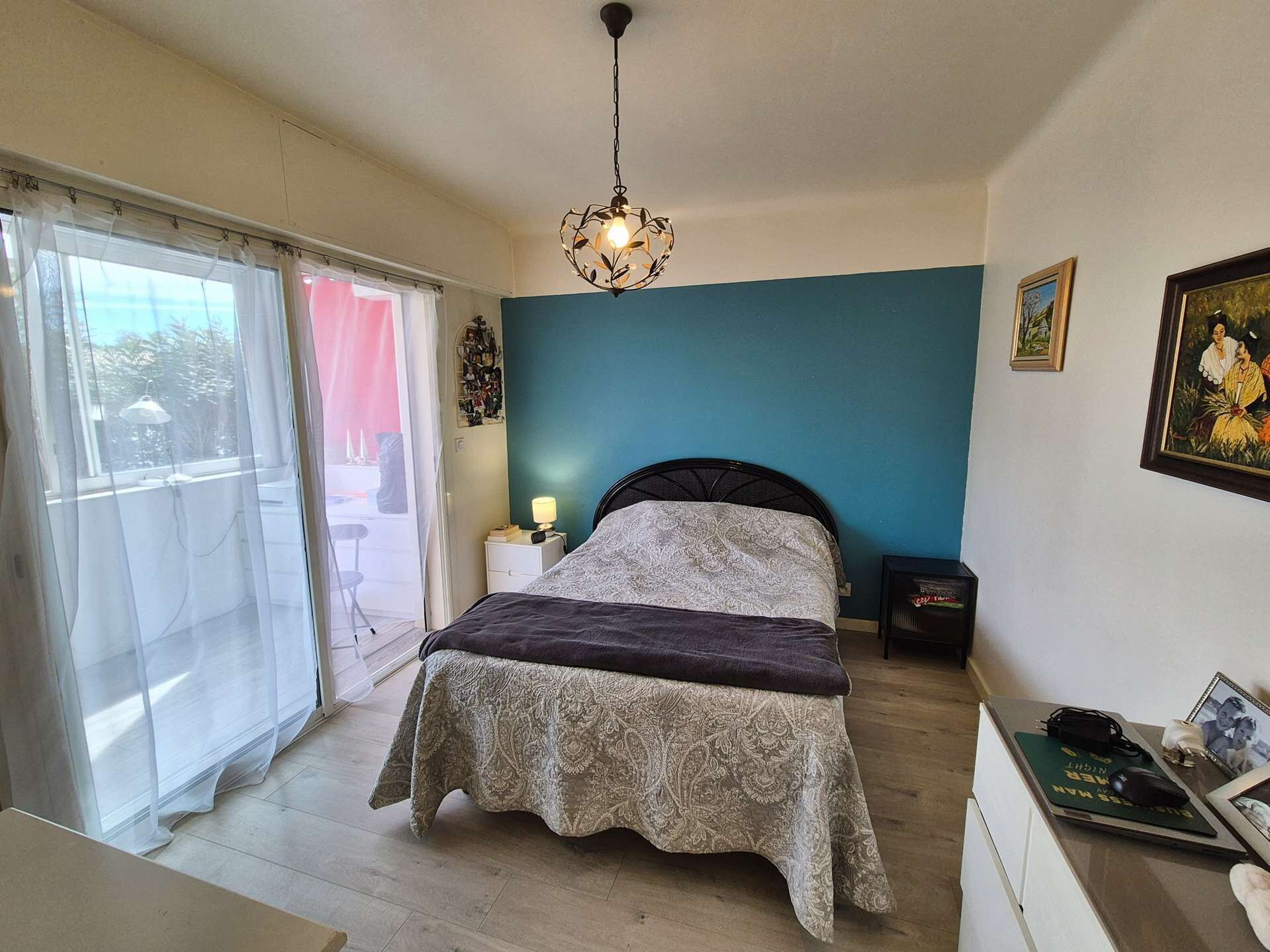 Een slaapkamer met een bed, nachtkastje, bureau, stoel, raam met dunne gordijnen en een plafondlamp.