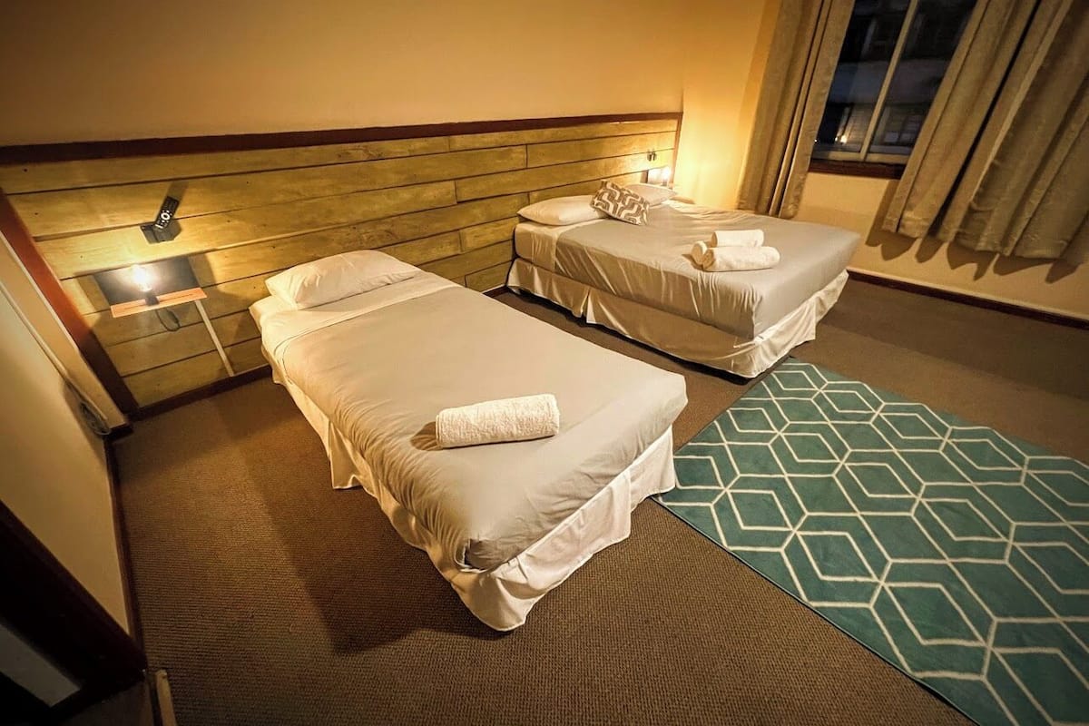 A hotel room with two beds, a wooden headboard, a green patterned rug, and a window with curtains.