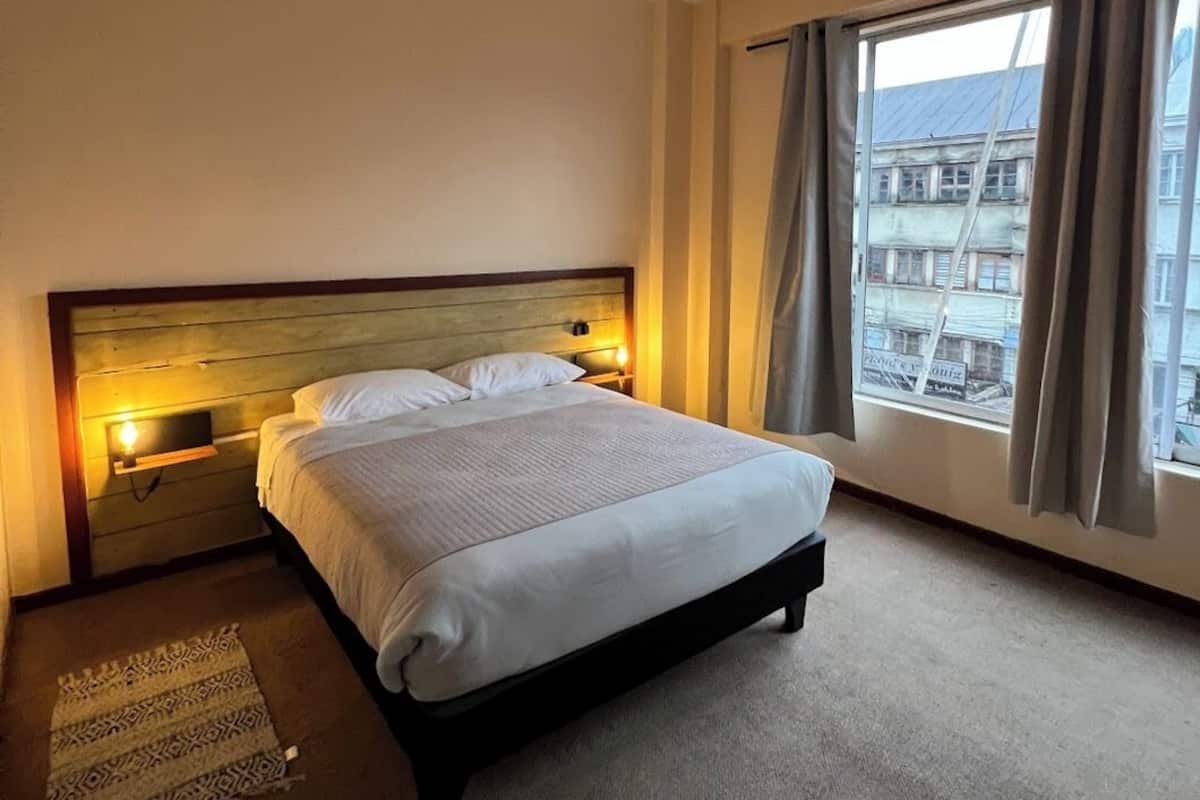 A hotel room with a large bed, bedside lamps, a view of buildings through the window, and a patterned rug on the floor.