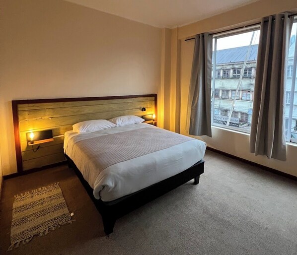 Une chambre d’hôtel avec un grand lit, des lampes de chevet, une vue sur les immeubles par la fenêtre et un tapis à motifs sur le sol.