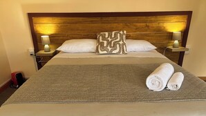 Habitación de hotel con cabecera de madera, dos lámparas de noche, almohada estampada, toalla enrollada y colcha con textura.