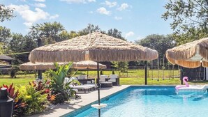 A swimming pool with thatched umbrellas, lounge chairs, and a flamingo float. - Barefoot Tiki Retreat -Inground Heated Pool, Hot Tub, Game Rm,Theater Rm,Firepit (Mason)