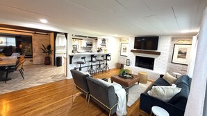 A modern living room with a fireplace, a kitchen area, and a dining space. - Barefoot Tiki Retreat -Inground Heated Pool, Hot Tub, Game Rm,Theater Rm,Firepit (Mason)