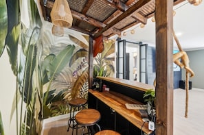 Een bar met een houten toonbank, krukken en een muurschildering van tropische planten.