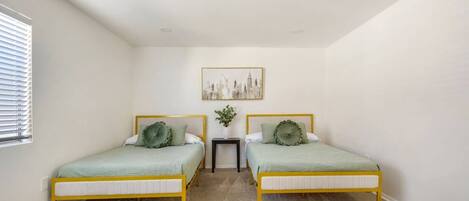 A bedroom with two beds, a small table, a plant, and a cityscape painting on the wall.