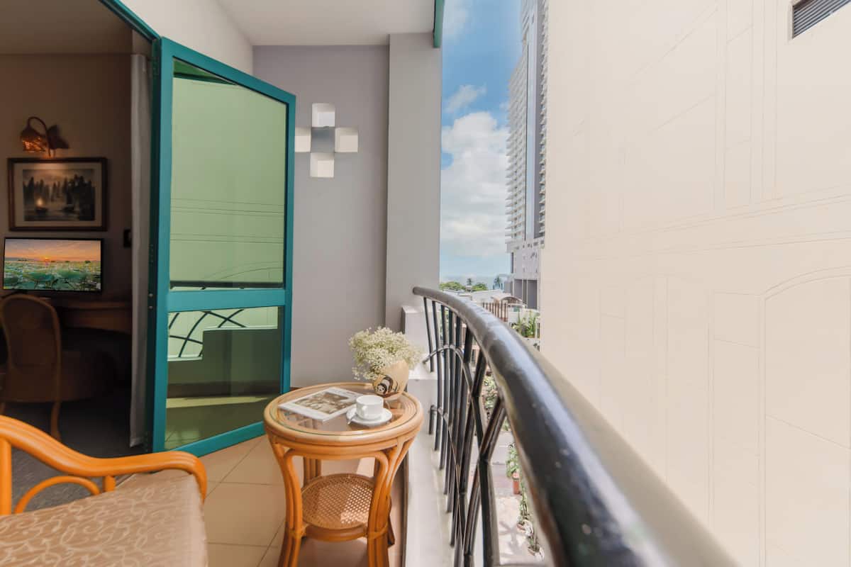 A balcony with a glass door, a wicker chair, a small round table with a cup and a vase, and a view of a tall building and a beach.