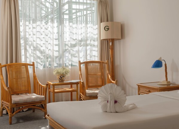 Une chambre d’hôtel avec un lit, deux fauteuils en osier, une petite table avec un vase de fleurs, une lampe et une fenêtre avec des rideaux.