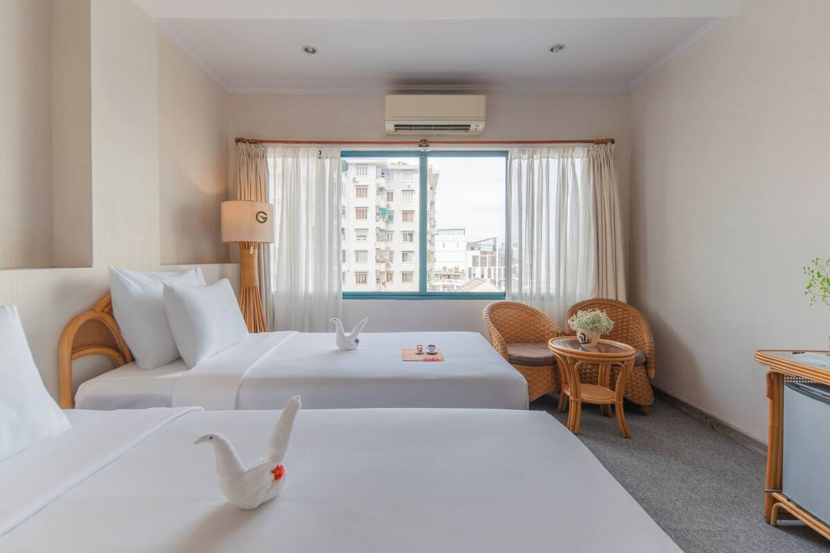 A hotel room with two beds, a small table with a teapot, a chair, and a window with curtains.