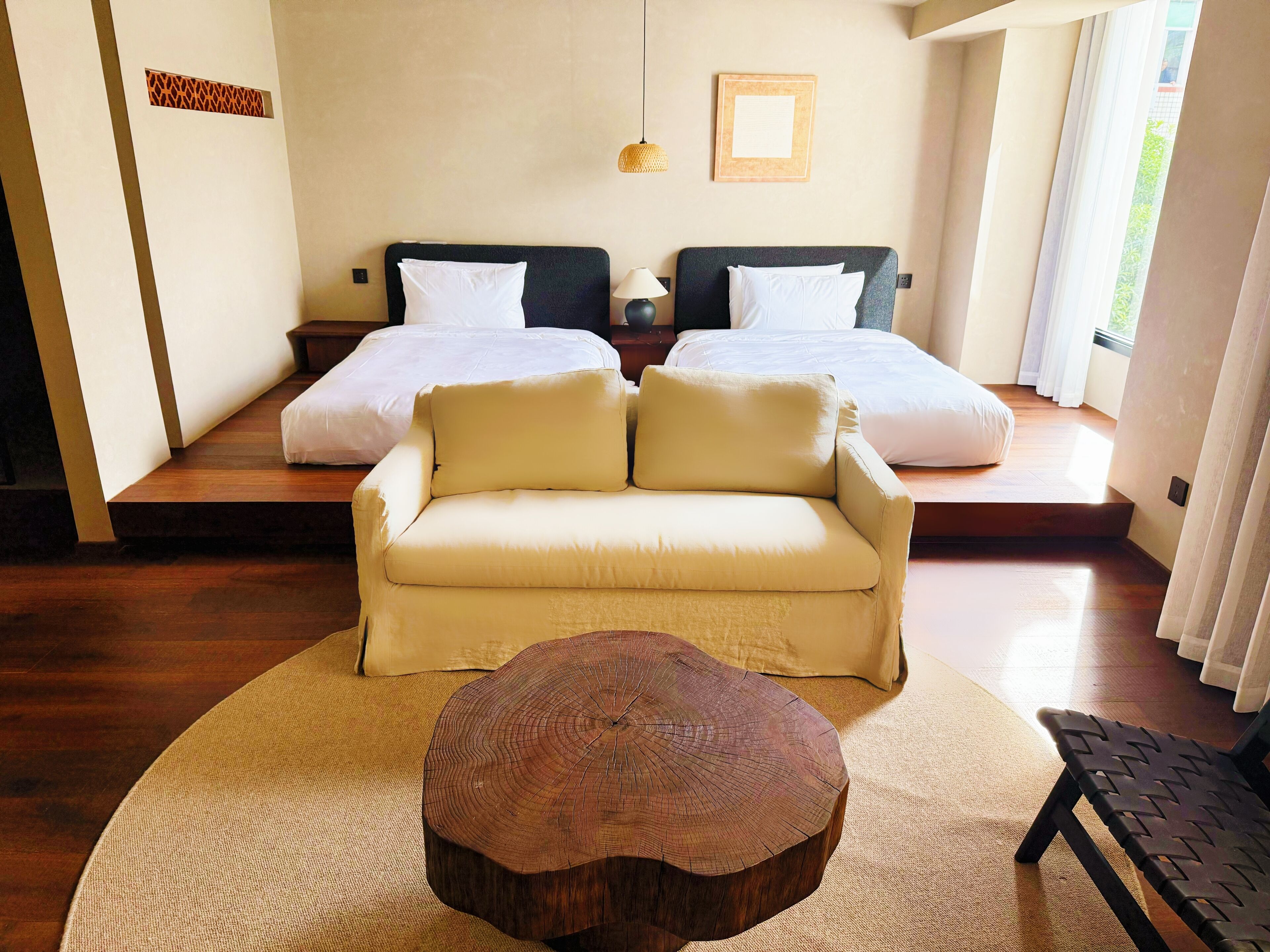 A room with a sofa, a round wooden coffee table, two beds with white bedding, a woven wall decoration, and a framed picture on the wall.