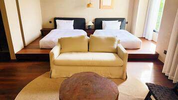 A room with a sofa, a round wooden coffee table, two beds with white bedding, a woven wall decoration, and a framed picture on the wall.