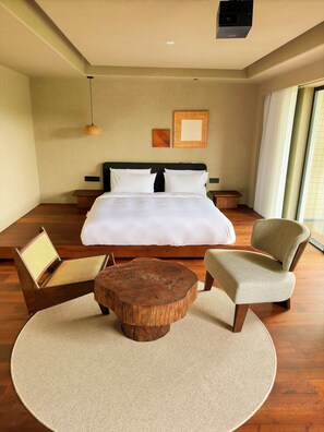 A bedroom with a bed, two chairs, a round wooden coffee table, a wall-mounted light, and a large window.