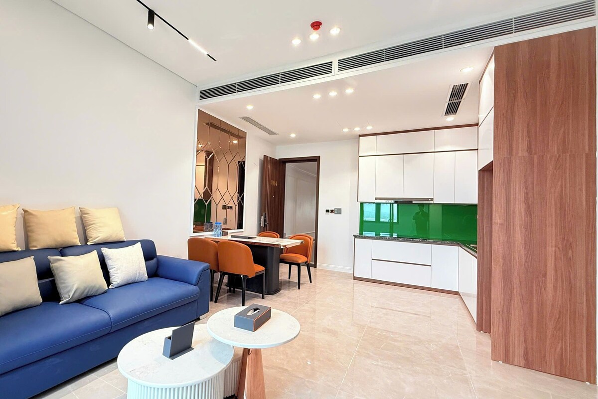 A modern living room with a blue sofa, a dining area with orange chairs, and a kitchen with white cabinets.