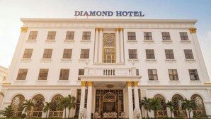 Un grand bâtiment blanc, sur lequel on peut lire l’inscription « DIAMOND HOTEL », avec une entrée imposante ornée de colonnes et de statues, ainsi que des fenêtres en arc de cercle.