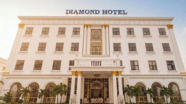 Un grand bâtiment blanc, sur lequel on peut lire l’inscription « DIAMOND HOTEL », avec une entrée imposante ornée de colonnes et de statues, ainsi que des fenêtres en arc de cercle.