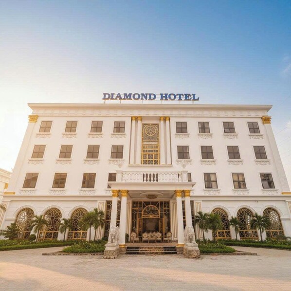 Un grande edificio bianco con insegna 'DIAMOND HOTEL', caratterizzato da un'imponente entrata con colonne e statue, e finestre ad arco.